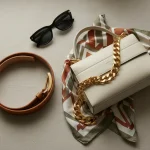 Accessori di moda donna per styling outfit: cintura in pelle, borsa strutturata, collana oro, occhiali da sole e sciarpa su sfondo neutro